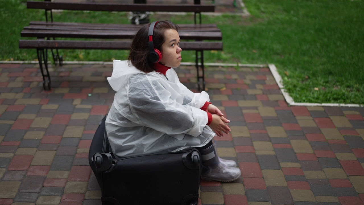 侧视图放松的小女人在雨衣和耳机听音乐微笑坐在手提箱在人行道上。愉快轻松的高加索小人儿旅人享受休闲。缓慢的运动。视频素材