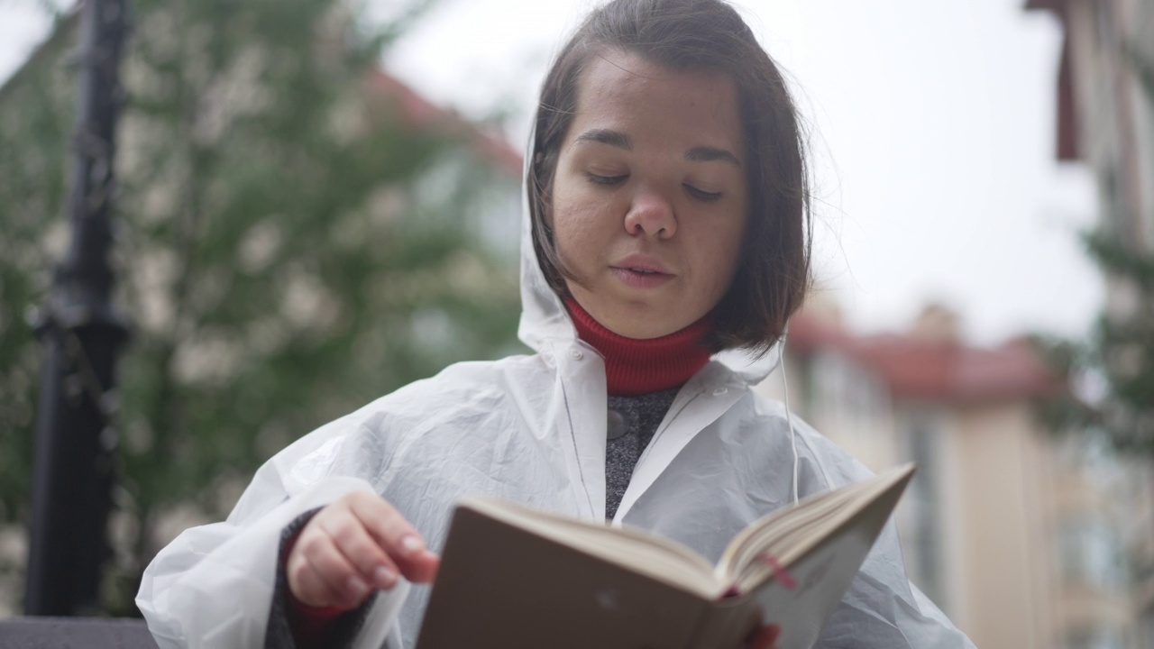 专注的白人小女人在雨衣阅读书在户外的慢动作微笑。自信快乐的小人物肖像享受在阴暗的周末在城市爱好。视频素材