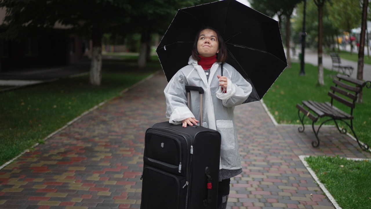 美丽的小女人带着大旅行袋站在户外，撑着雨伞向上看。快乐自信的高加索小人儿在雨天的城市旅行。缓慢的运动。视频素材