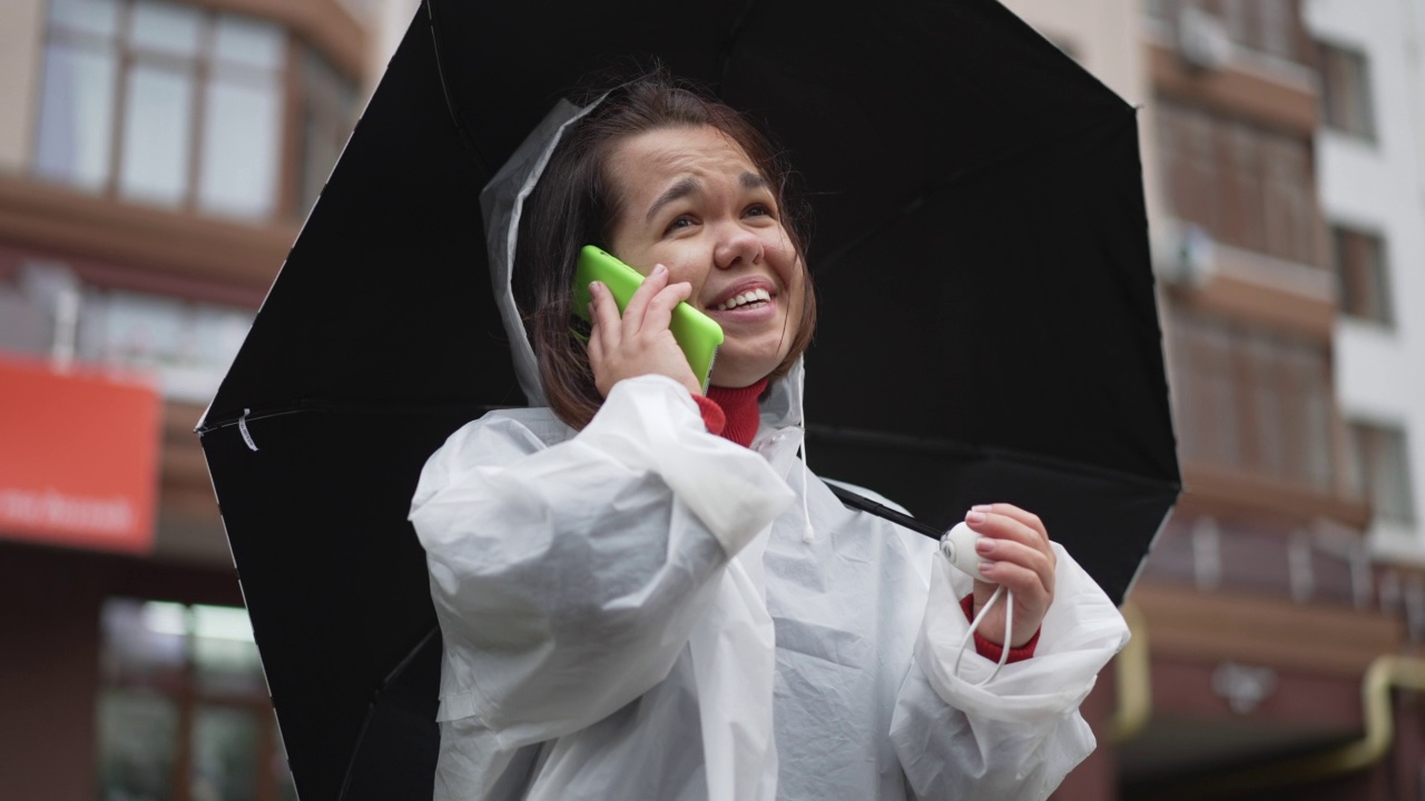 开朗的小妇人在雨天站在户外笑着讲电话。穿着雨衣、打着雨伞、在城市中使用智能手机的快乐白人小人物的肖像。视频素材