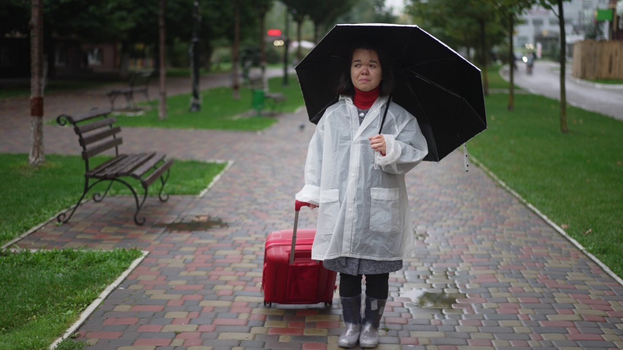 崩溃沮丧的小女人站在城市街道上哭泣，带着雨伞和旅行袋。在阴天的户外，沮丧悲伤的白人小个子游客的广角肖像。缓慢的运动。视频素材