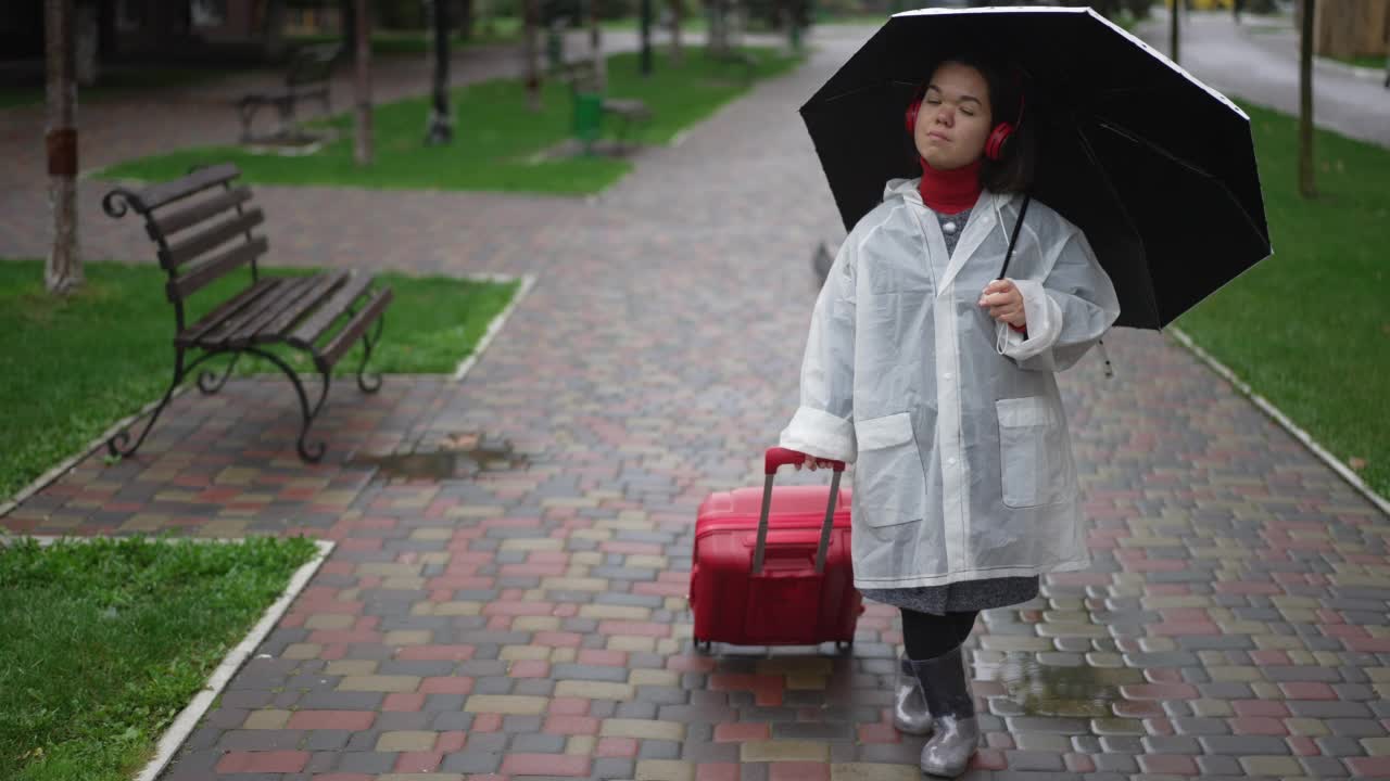 年轻的小女子穿着雨衣和胶靴，带着雨伞和旅行包站在城市的户外。白种人小个子在阴天旅行的宽镜头肖像。视频素材