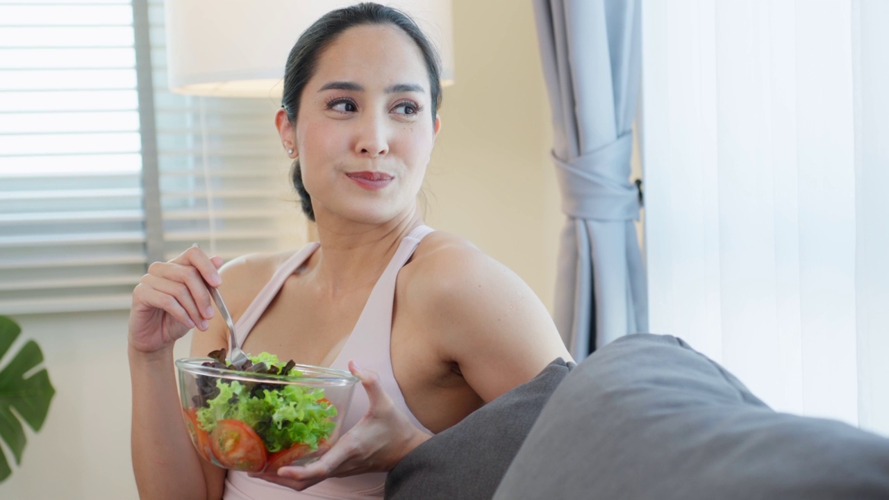 亚洲有吸引力的运动妇女拿着沙拉碗和吃蔬菜。漂亮的运动女孩穿着运动服，运动后喜欢在家里吃干净的蔬菜，以保持健康。饮食与健康食品理念。视频素材