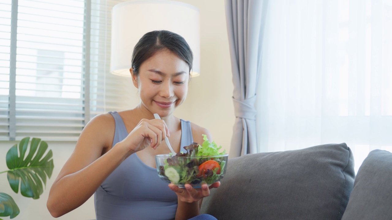 亚洲有吸引力的运动妇女拿着沙拉碗和吃蔬菜。漂亮的运动女孩穿着运动服，运动后喜欢在家里吃干净的蔬菜，以保持健康。饮食与健康食品理念。视频素材