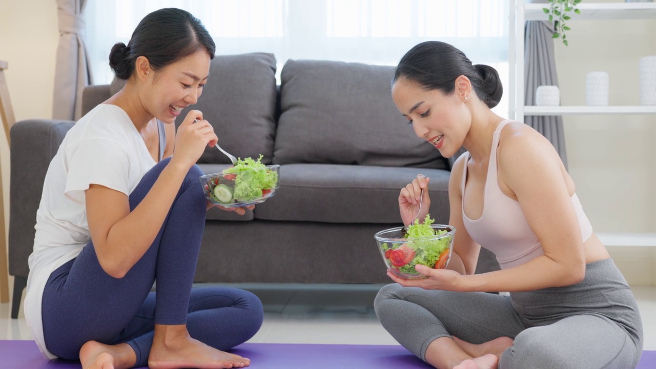 亚洲吸引人的两个运动女人拿着沙拉碗和吃蔬菜。漂亮的运动女友穿着运动服，喜欢在家里吃干净的蔬菜，运动后一起带走。视频素材