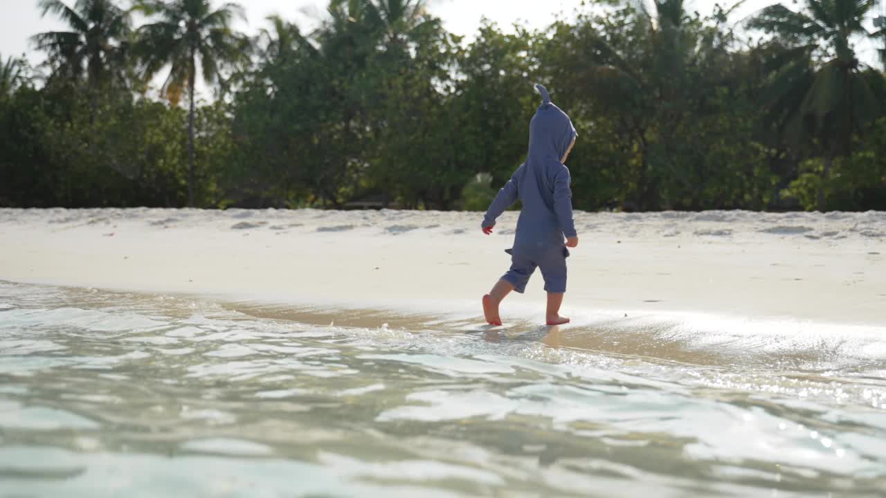 一个穿着鲨鱼服装的孩子沿着海洋的热带海滩奔跑。视频素材