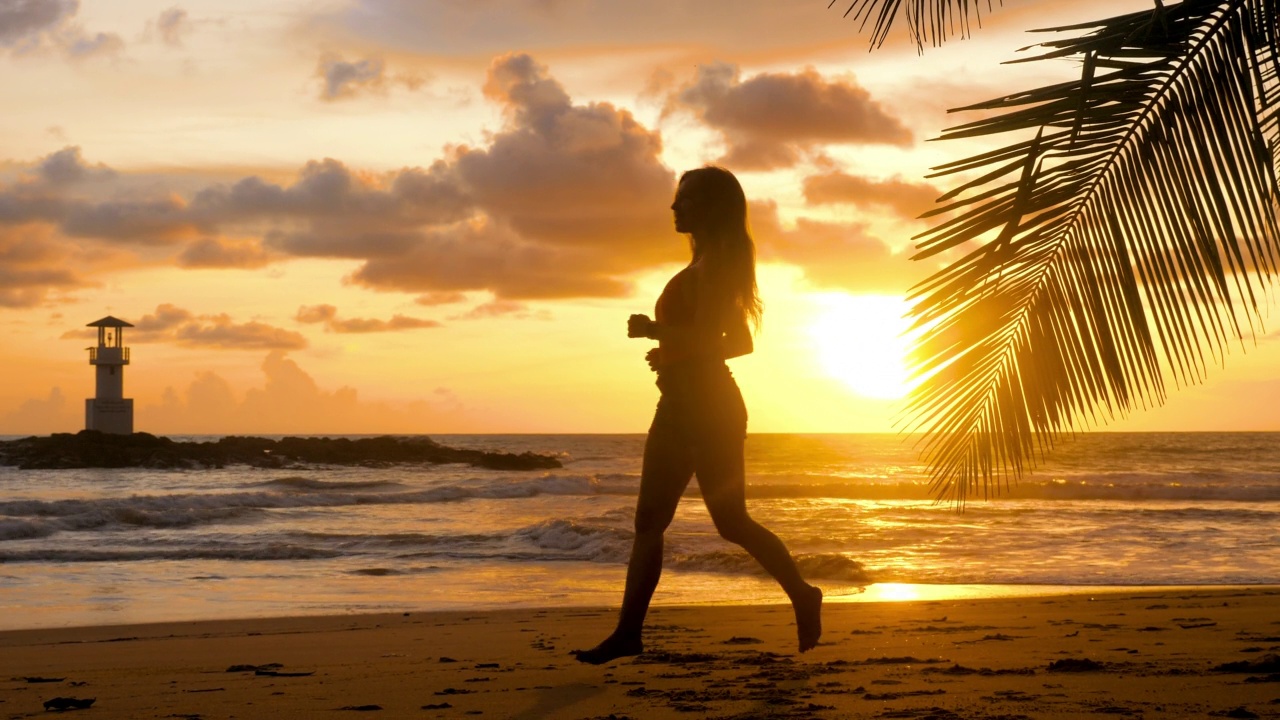 年轻女子锻炼跑步赤脚在海滩上与日落海洋背景。夏日海景，健康积极的生活方式，户外慢跑运动理念。女性运动员廓形有氧训练视频素材