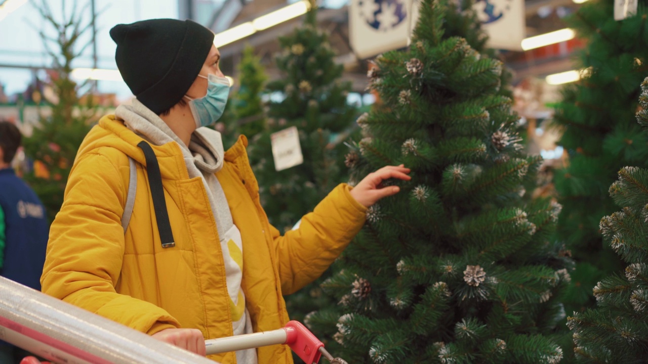 一名女子在市场上挑选圣诞树。身穿黄色冬装、戴着医用防护口罩的白人女性在购物中心触摸人造圣诞树视频素材