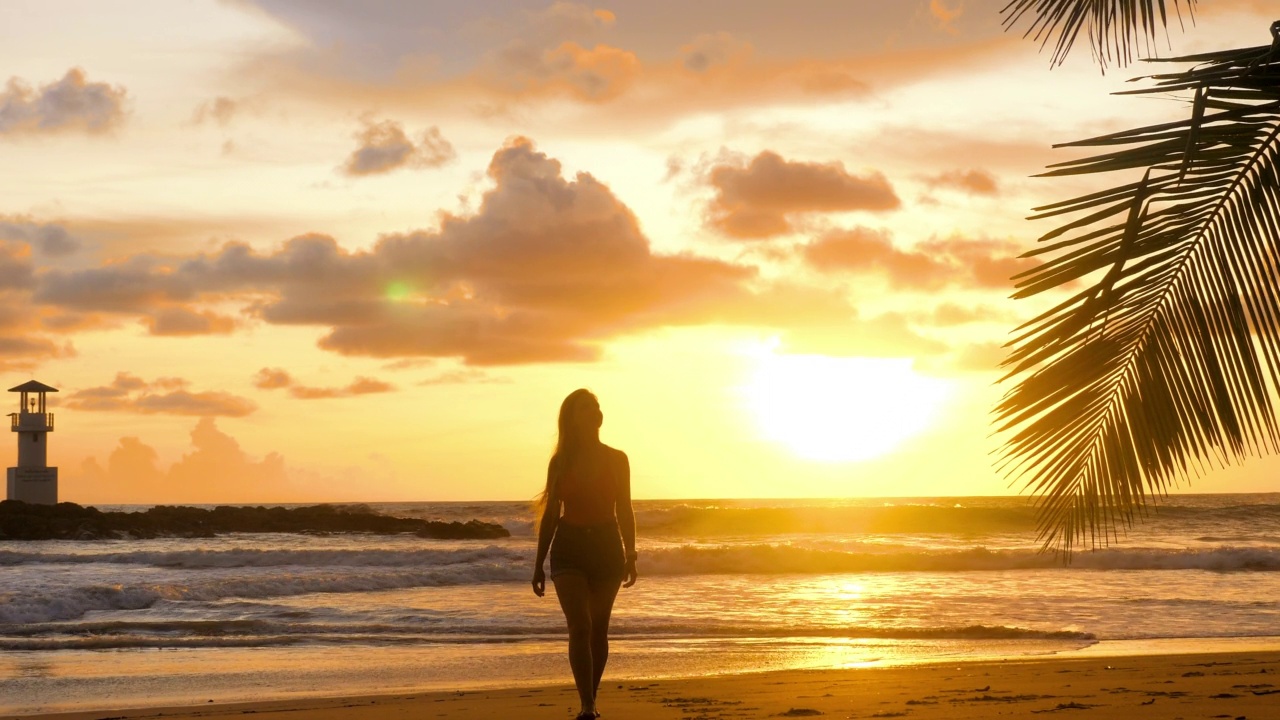 年轻的女人走在浪漫的金色夕阳灯光与海浪背景。快乐女孩享受暑假假期在海洋。在沙滩上赤脚走在海边的女游客。慢动作视频素材