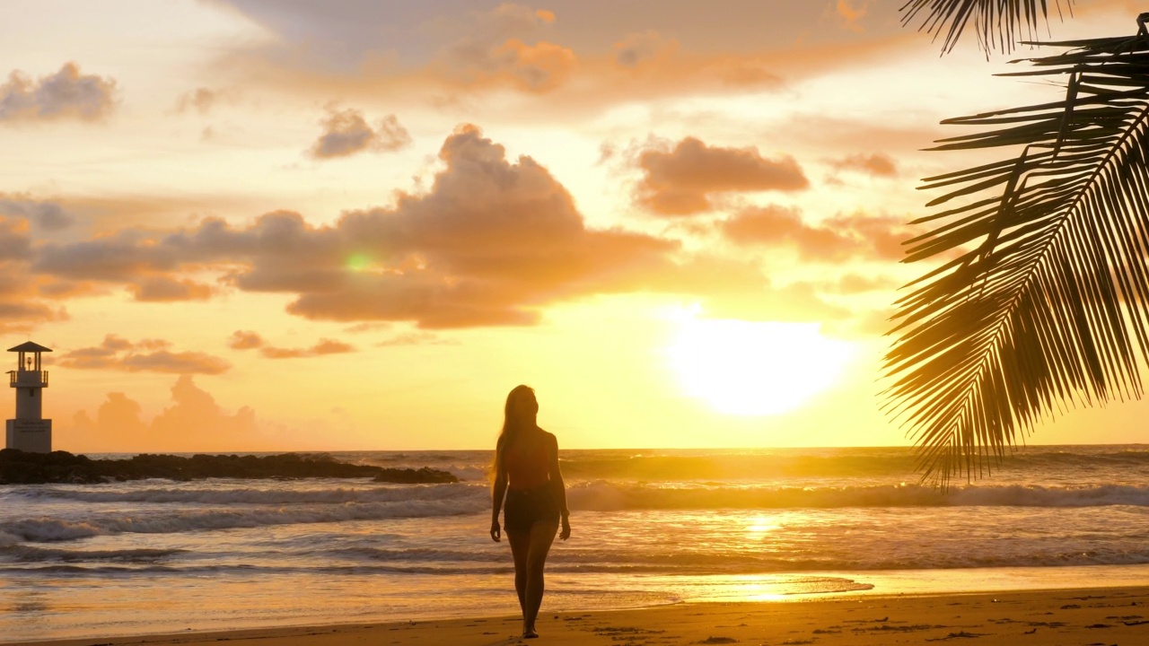 年轻的女人走在浪漫的金色夕阳灯光与海浪背景。在沙滩上赤脚走在海边的女游客。快乐女孩享受暑假假期在海洋。慢动作视频素材