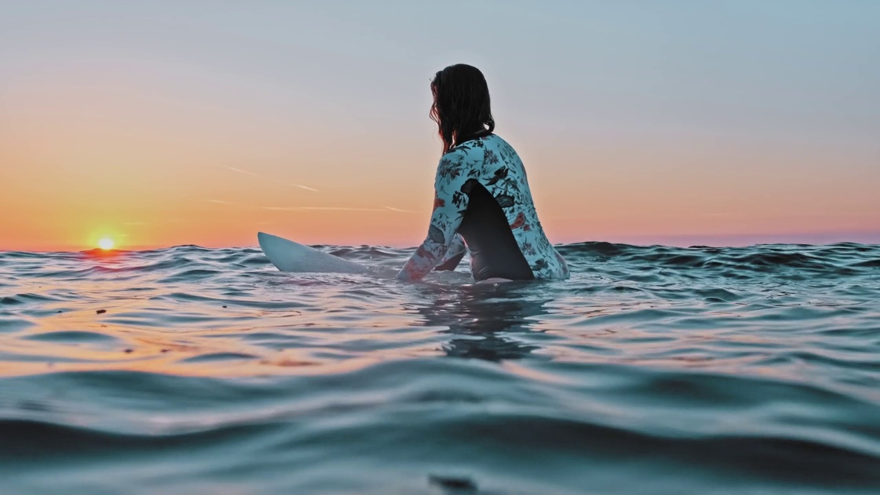 女冲浪者在长袖泳衣坐在冲浪板上漂浮在平静的波浪大海看地平线金色的日落视频素材