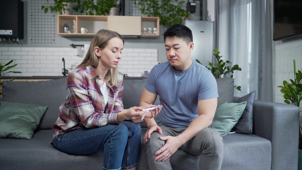 一对夫妇在怀孕测试结果阴性后伤心难过。忧心忡忡的年轻女性和亚洲男性坐在家里的沙发上，手里拿着棍子视频素材