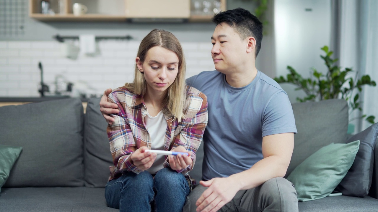 一对夫妇在怀孕测试结果阴性后伤心难过。忧心忡忡的年轻女性和亚洲男性坐在家里的沙发上，手里拿着棍子视频素材