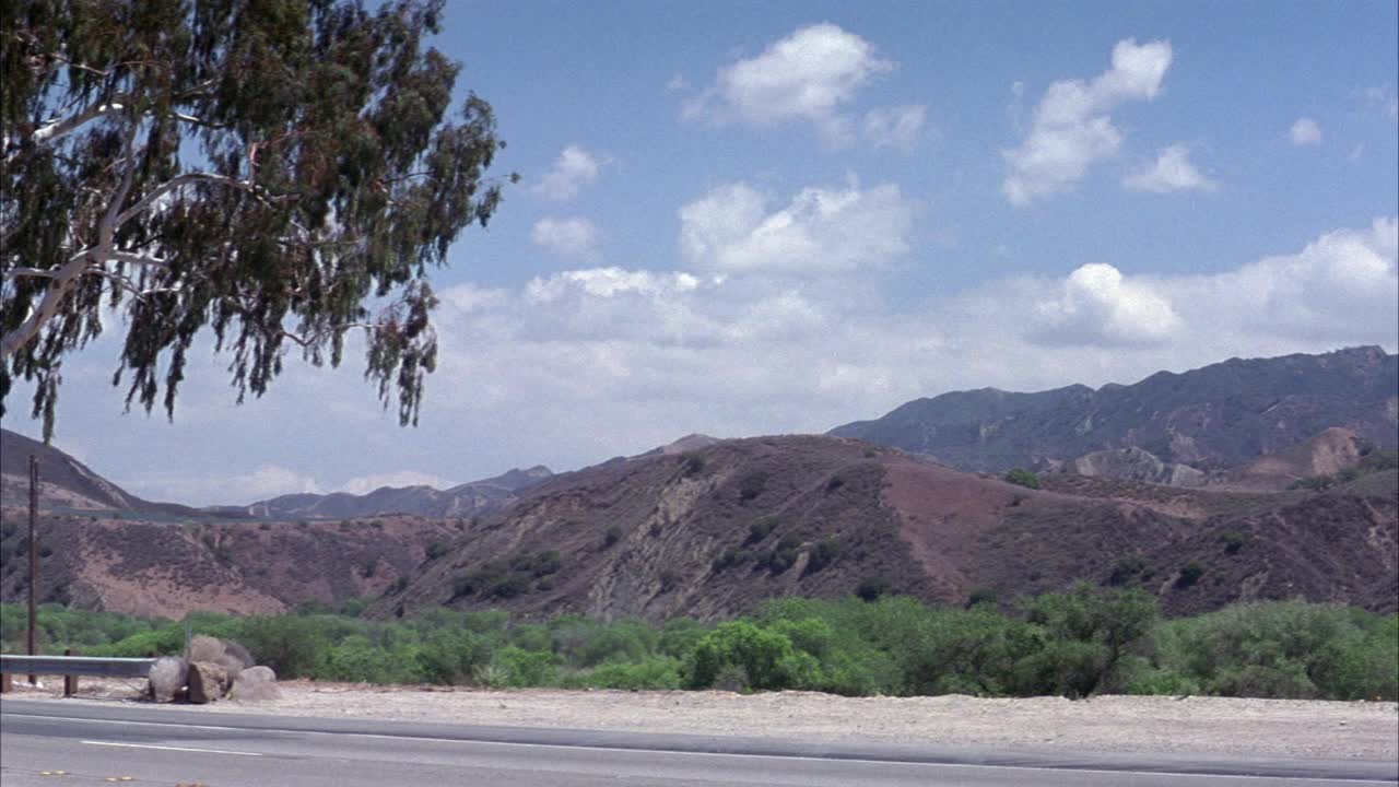 沙漠公路中角。看到路边的路边。在背景中看到沙漠的山丘或山脉。看到绿色的树叶和灌木。看到黑色福特astro厢式货车从左边突然转向，在路边刹车。前景见土云视频素材