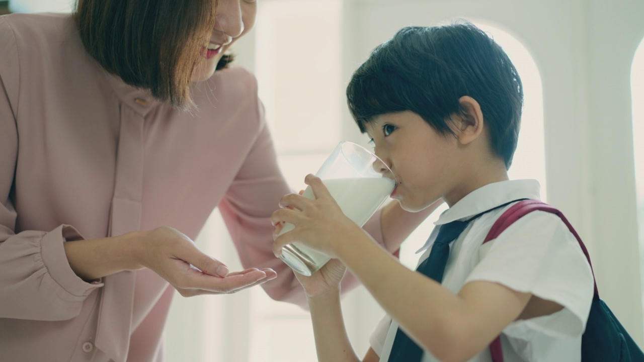 美丽的亚洲妈妈正在鼓励穿着校服和背包的小儿子在早上上学前喝鲜奶，以保持孩子健康的生活方式。小男孩在家用玻璃杯喝鲜奶。视频素材