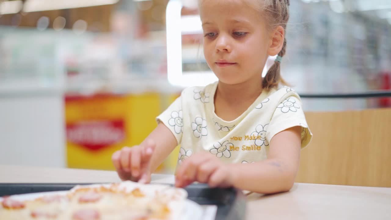 孩子坐在超市食品广场的桌子上吃披萨。小女孩在室内享受快餐营养。饥饿的孩子在吃披萨。肖像拍摄。视频素材