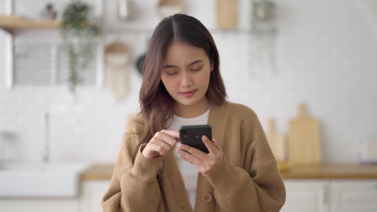 快乐的年轻亚洲女人在家里放松。亚洲女性站在厨房柜台，使用手机视频素材