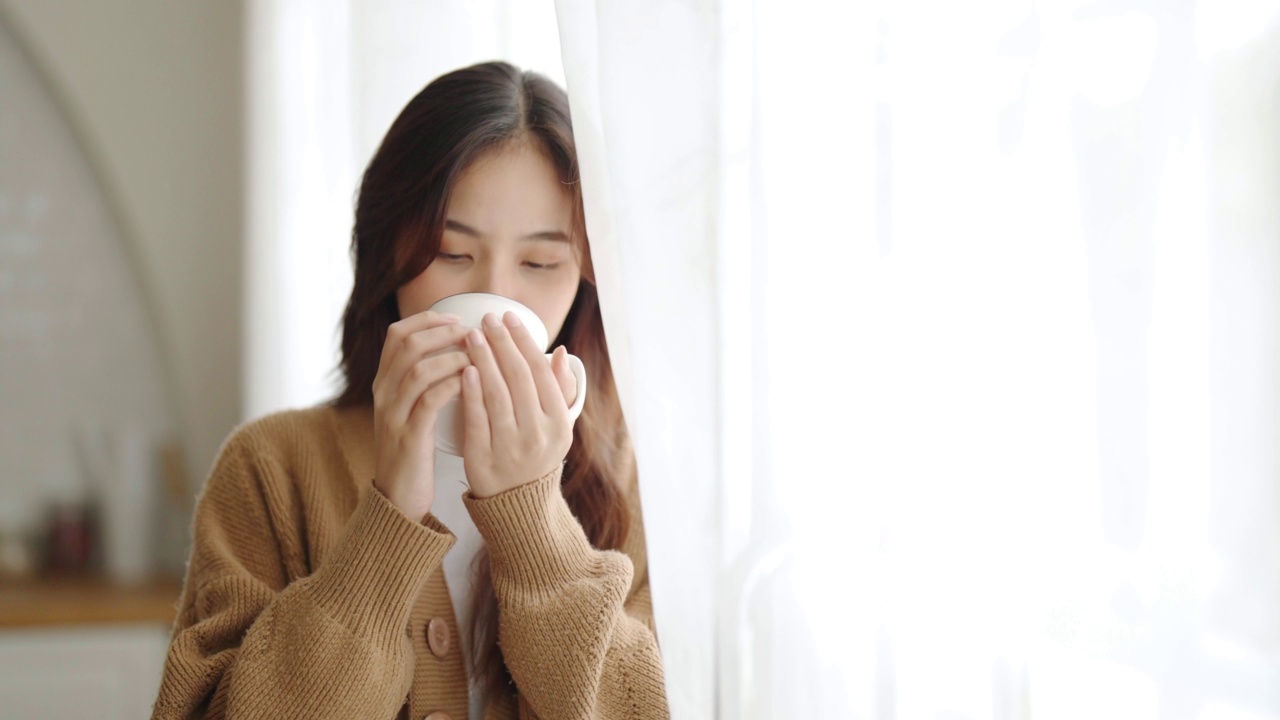 年轻的亚洲女性站在窗边，拿着杯子在家里的客厅里。亚洲女性早上醒来后喝牛奶或茶视频素材