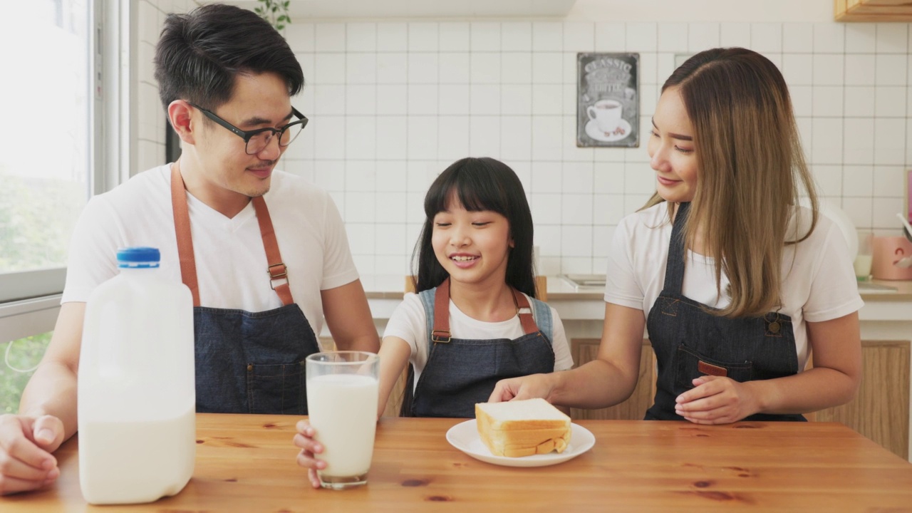 亚洲家庭在舒适的厨房享受早餐，小女孩女儿坐在桌子上，喝牛奶微笑在早上。幸福的家庭在客厅。视频素材