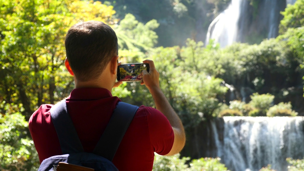 快乐的男性游客与背包旅行。漫游癖、旅游概念。年轻的旅行者用智能手机拍下了令人惊叹的瀑布和热带自然的风景视频，并在社交媒体上发布视频素材