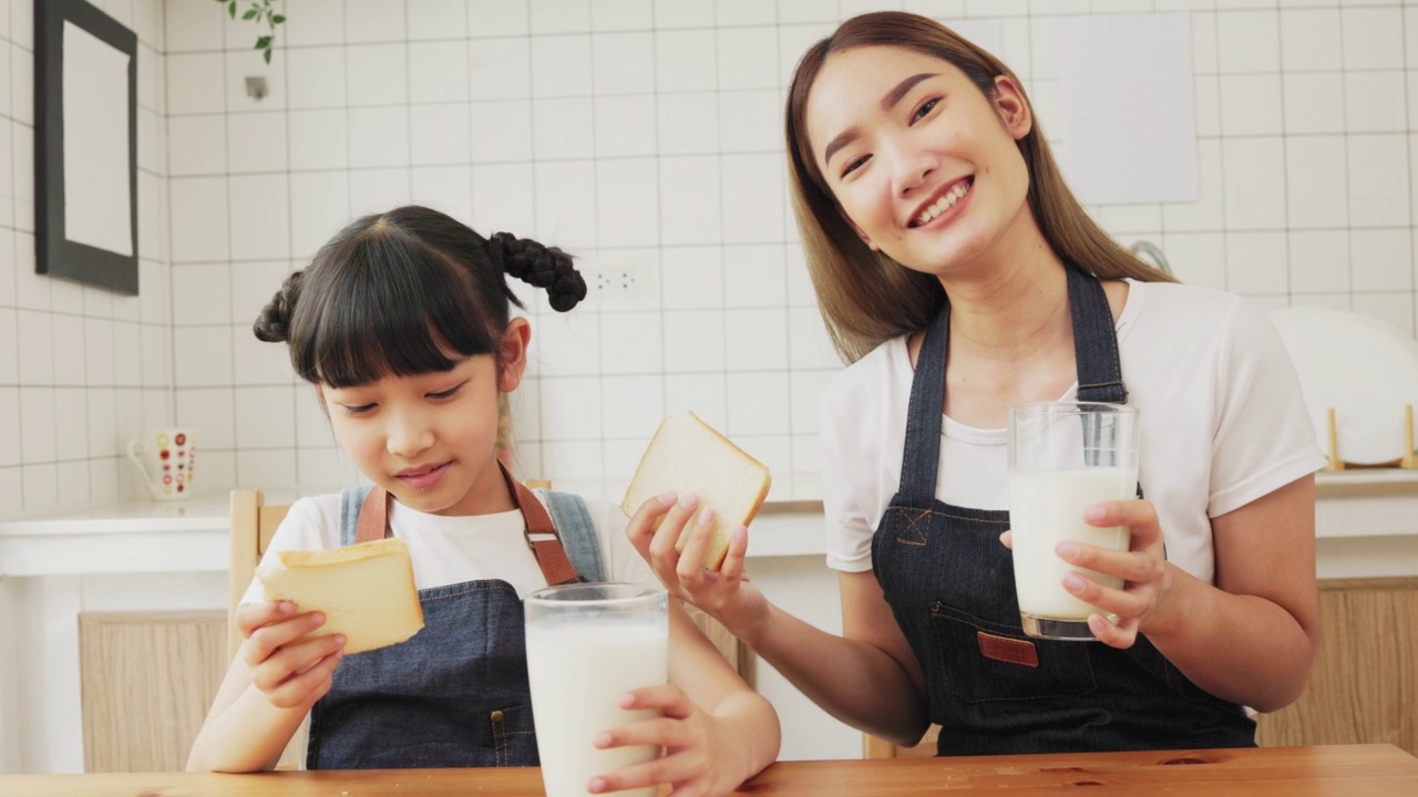 亚洲家庭在客厅享用早餐。小女孩女儿坐在桌子上，和微笑的爸爸妈妈在早上喝牛奶。家庭幸福。视频素材