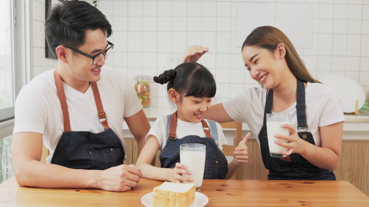 亚洲家庭在舒适的厨房享受早餐，小女孩女儿坐在桌子上，喝牛奶微笑在早上。幸福的家庭在客厅。视频素材