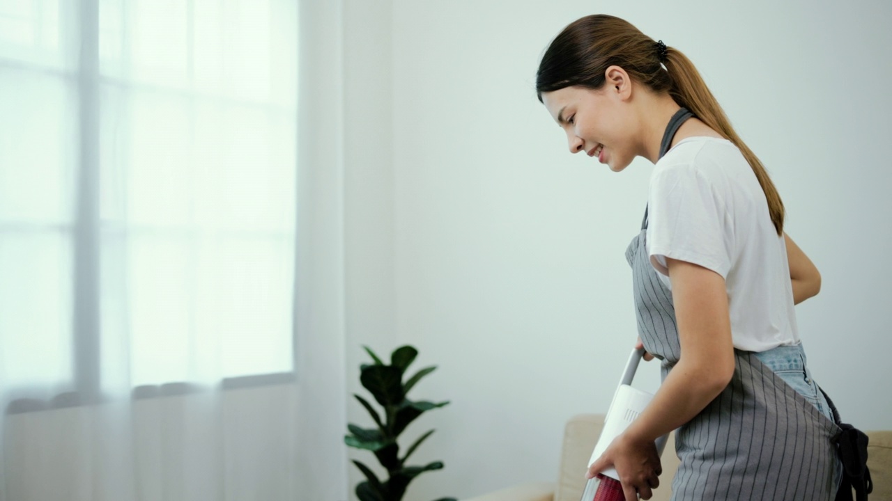 美丽的家庭主妇穿着围裙，用吸尘器打扫客厅。年轻女子很乐意打扫家里。女服务员清洁服务。视频素材