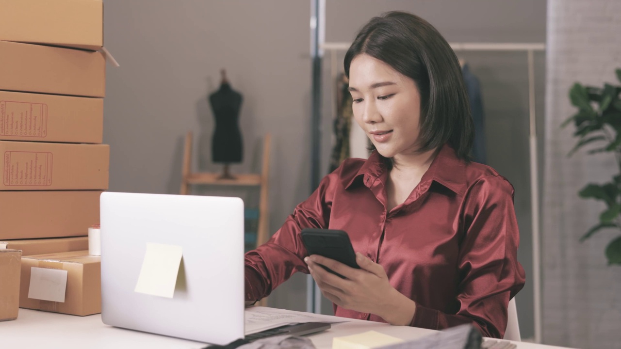 一名年轻的亚洲女子微笑着使用手机，身边有笔记本电脑和纸板盒，准备她的网上生意视频素材