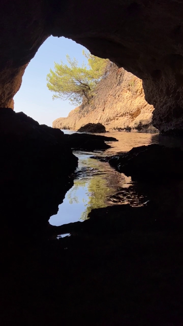 通过一个黑暗的山洞的一个洞，可以看到海水和岩石，还有松树。视频素材