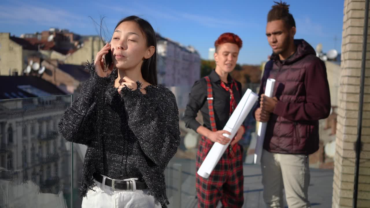 美丽的亚洲年轻女子的肖像笑着讲电话在慢动作模糊的白种人和非洲裔美国承包商的背景。自信的女士在阳光下的建筑露台。视频素材