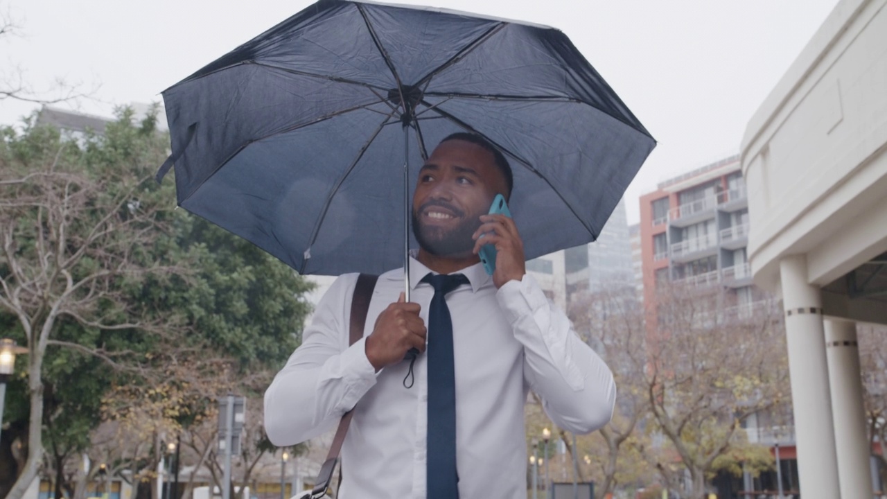 英俊的年轻商人在雨中打着伞走在城市里打电话。在冬天通勤上班。在电话中提供保险的拉丁公司保险代理人视频素材