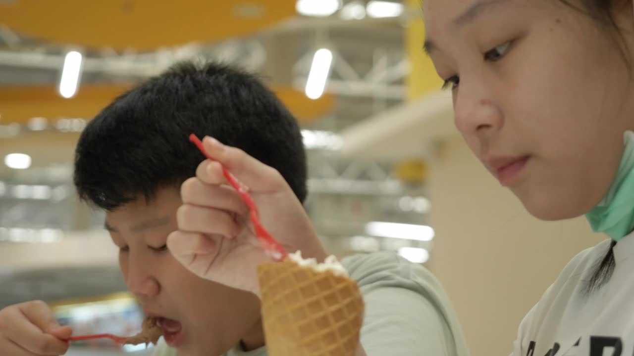 亚洲小孩在美食广场一起吃冰淇淋，男孩在玩手机游戏。视频素材