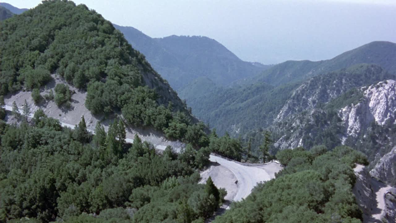 褐色和棕色的凯迪拉克沿着山边的道路行驶的航拍。看到常青树覆盖山脉和周围的道路视频素材