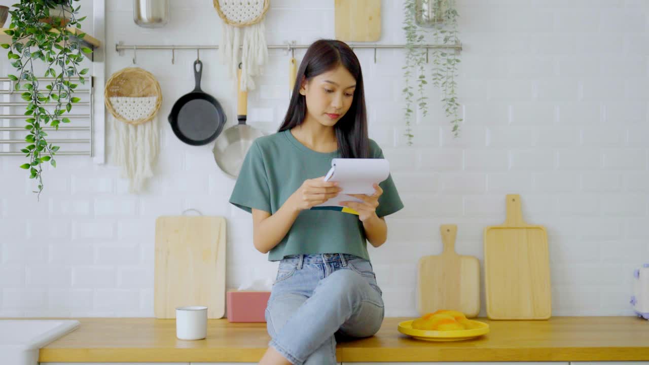 快乐的年轻亚洲女人坐在家里的厨房柜台上。女性读书笔记烹饪视频素材