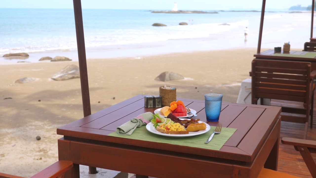 在阳光明媚的夏日，泰国普吉岛海滩上的户外咖啡馆与食物的桌子。旅行，度假，度假的概念。热带国家的海滨餐厅，绿松石海浪的背景视频素材