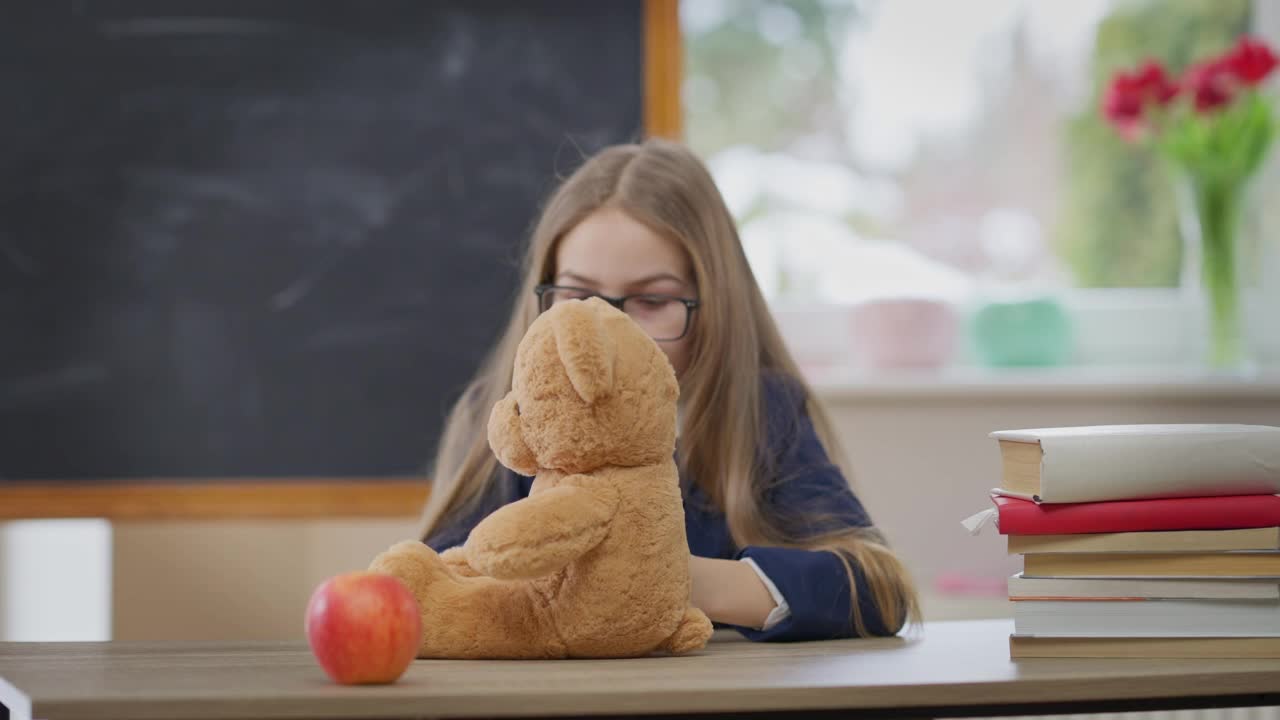 机架焦点从泰迪熊到学生女孩手势大拇指向上微笑看着相机。十几岁的白人女孩的肖像在课桌与玩具在教室。教育和信心。视频素材