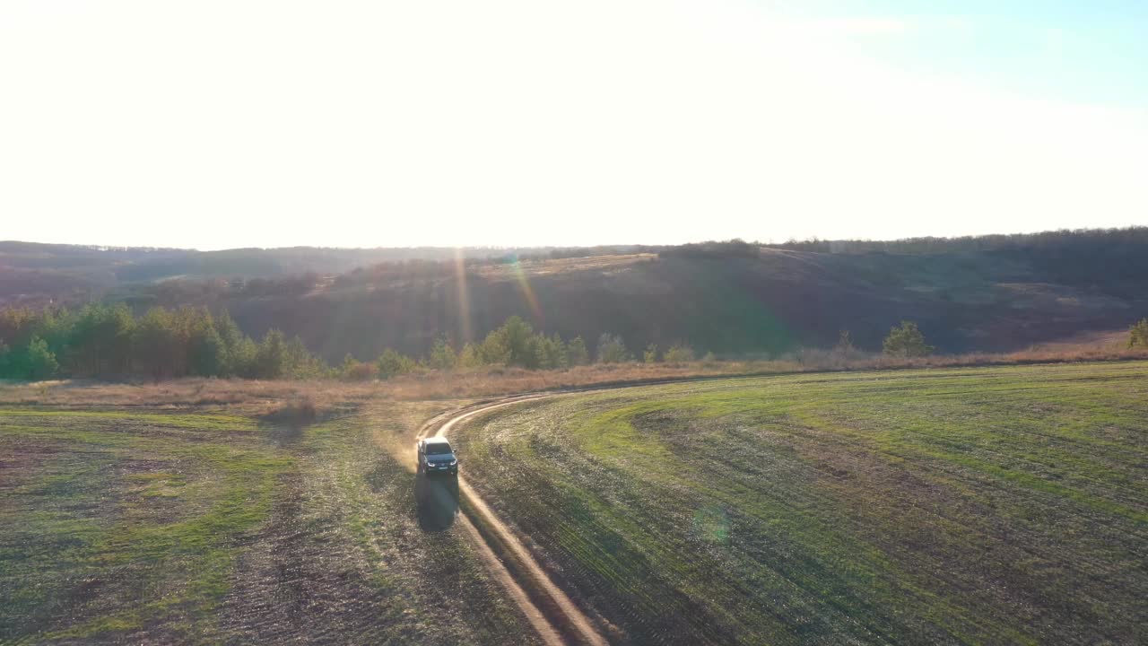 以阳光为背景，沿着田野附近的道路行驶的皮卡。在乡间行驶的黑色汽车。初秋时，在靠近草地的乡间小路上行驶的越野车。空中拍摄视频素材