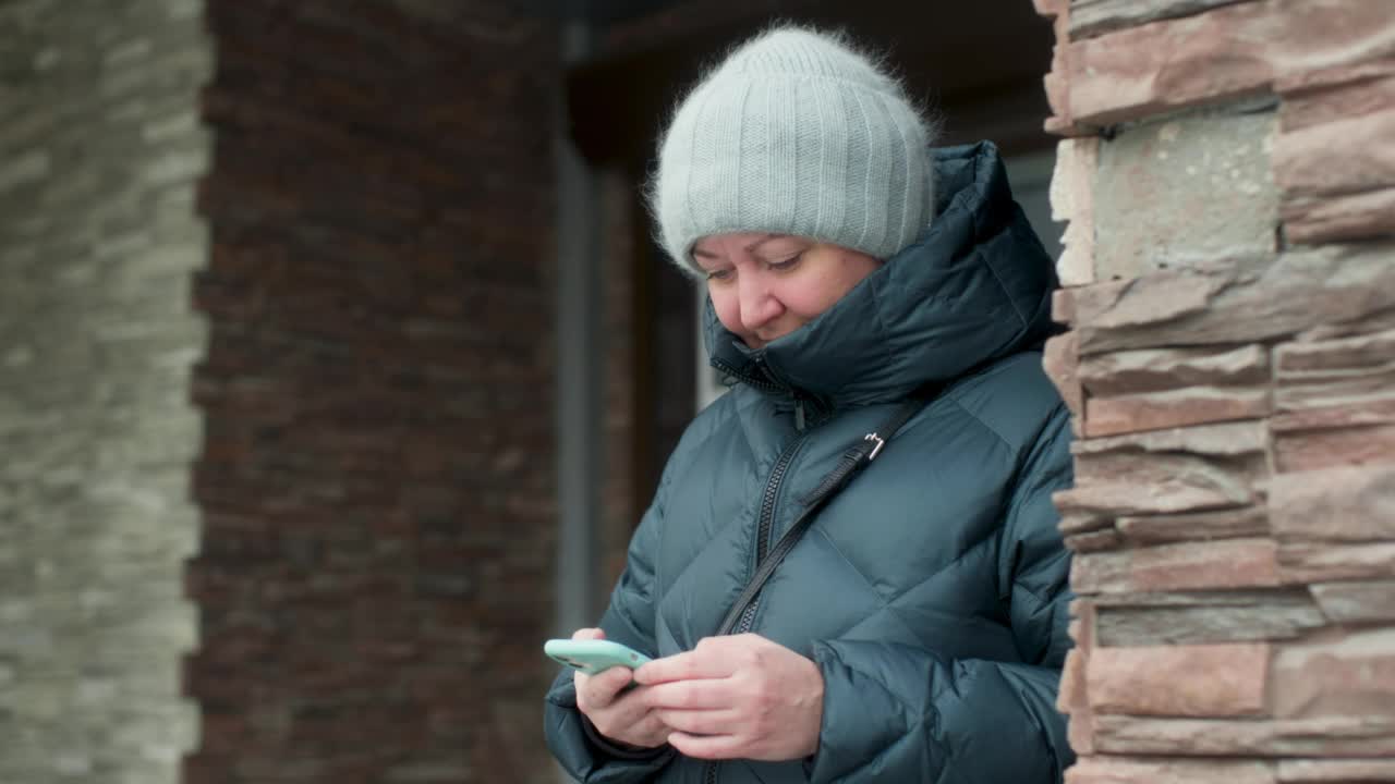 穿着保暖外套的白人女性，在寒冷的秋冬，在室外靠近石头砖墙的地方使用手机，用手指看手机和触摸屏幕。视频素材