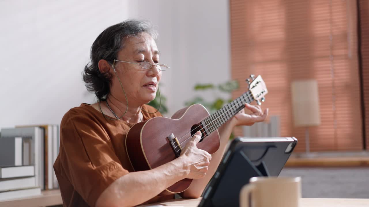 亚洲成熟的老年女性戴着眼镜练习和学习弹吉他通过观看在线数字平板电脑视频素材