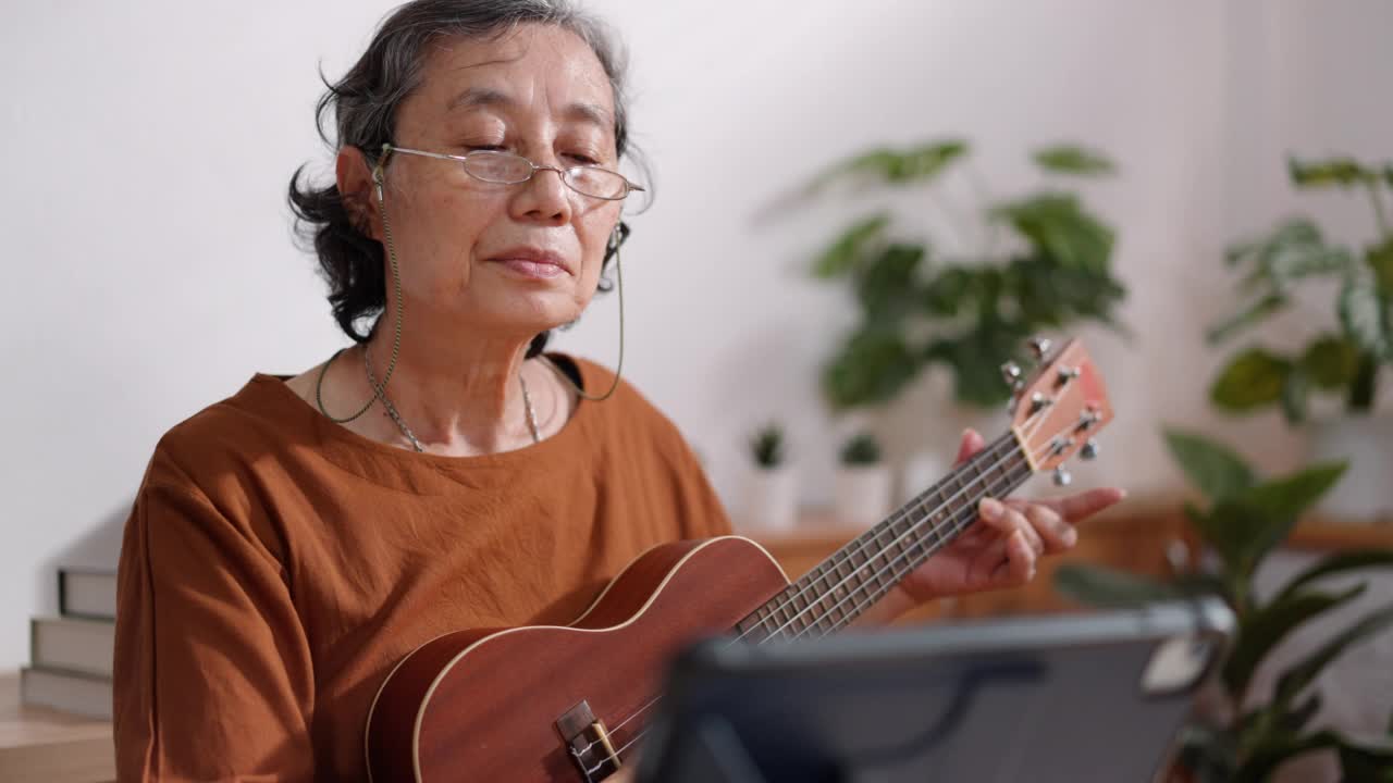 亚洲成熟的老年女性戴着眼镜练习和学习弹吉他通过观看在线数字平板电脑视频素材