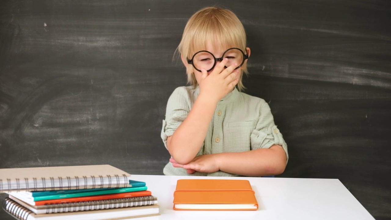 回到学校。快乐微笑的小学生戴着圆眼镜坐在课桌前。教室里有个孩子在黑板上举手。小男孩，上小学的。秋天的第一天。视频素材