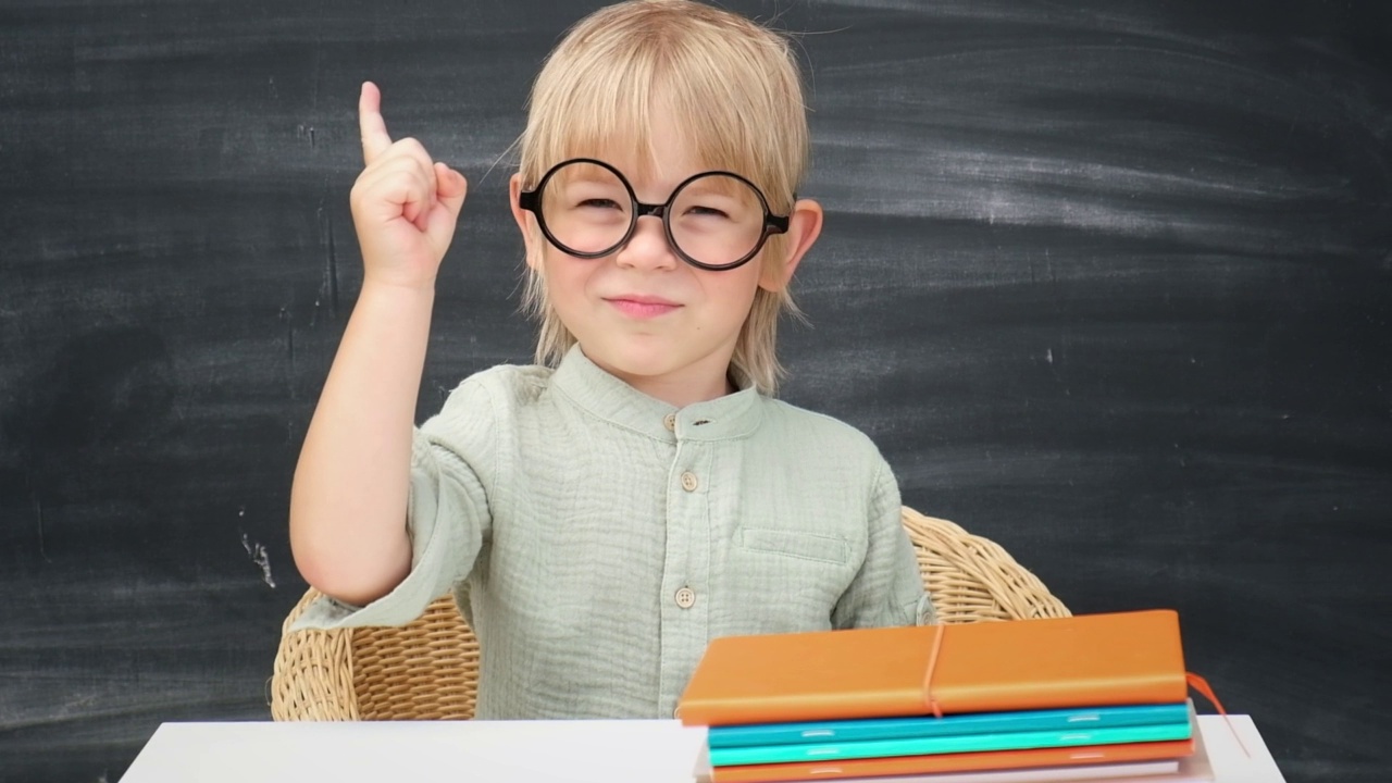 回到学校。快乐微笑的小学生戴着圆眼镜坐在课桌前。孩子们在教室里用黑板做背景。小男孩，上小学的。秋天的第一天视频素材