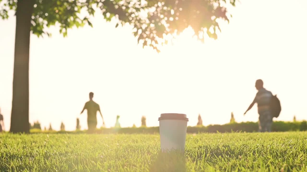 白色的纸咖啡茶杯，黑色的塑料盖子，站在绿色的草地在公园的夕阳。模糊的行人剪影在背景上。热饮外带，外卖咖啡茶视频素材