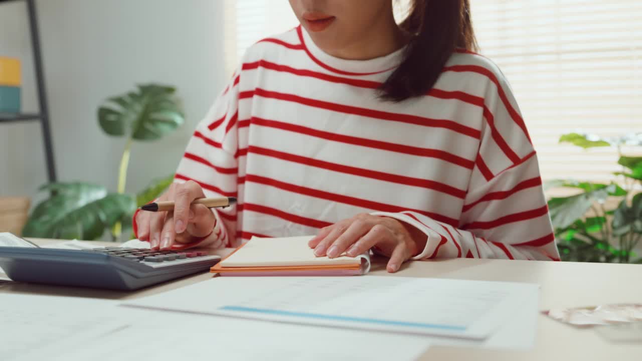 特写年轻的亚洲女人与毛衣坐在前台与笔记本电脑使用计算器计算水电费帐单检查信用卡收据每月的费用账单在家里。视频素材