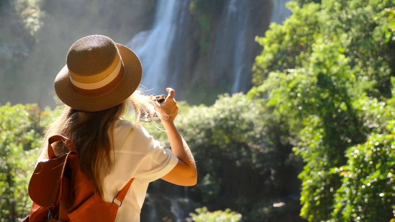 用相机拍摄热带瀑布的女人的背影。激励千禧一代旅行博主在旅途中。快乐的年轻旅行者女孩在暑假。旅游和冒险的概念。视频素材