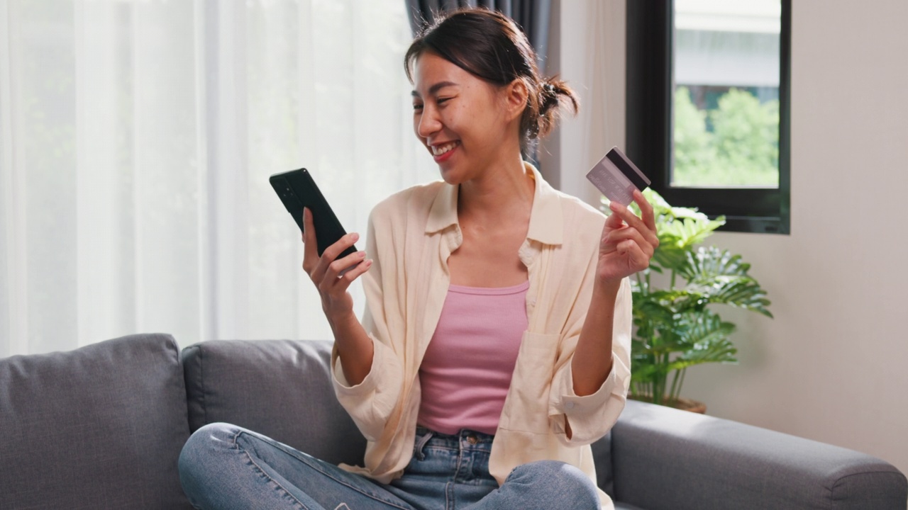 年轻的亚洲女女孩穿着休闲坐在沙发上，抱着智能手机和信用卡感觉兴奋和幸福的网上购物成功支付在客厅在家里。视频素材