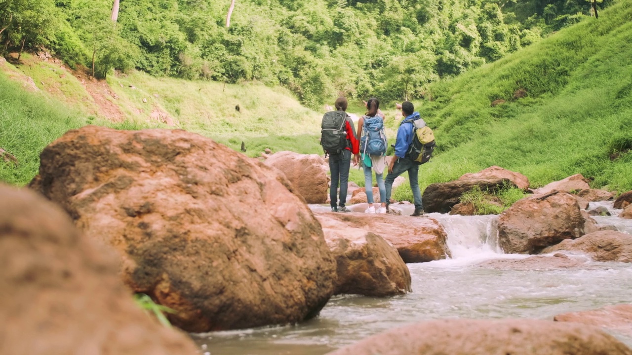 一群背着背包的徒步旅行者站在绿色山谷中一条小溪的岩石上。视频素材