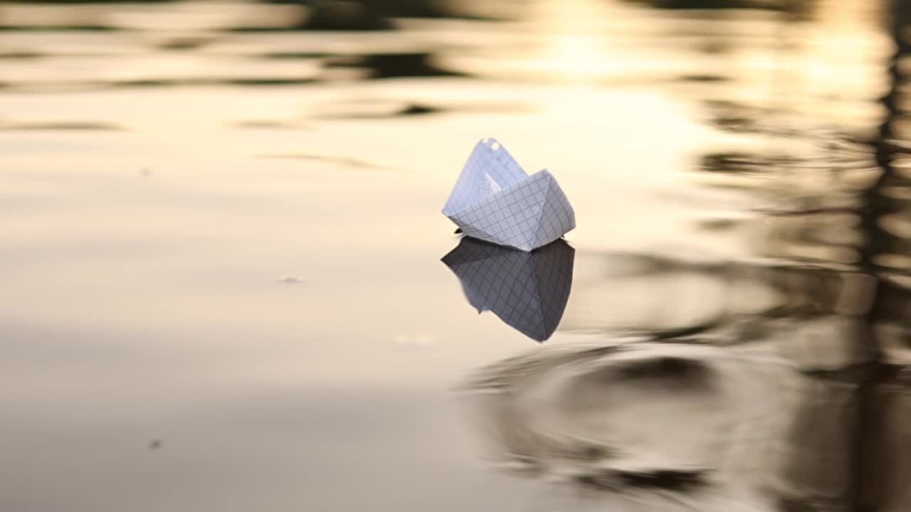 在美丽的夕阳下，一只纸船漂浮在水面的波浪上。折纸船。梦想、未来、童年、自由或希望的概念。缓慢的运动。高质量的全高清镜头。视频素材