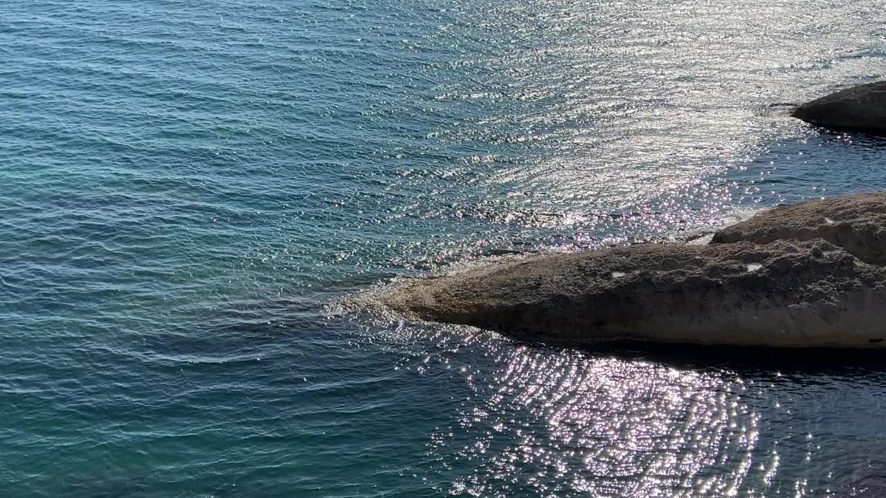 美丽的绿松石海景。风景多岩石的海岸线。波光粼粼的清澈海水。海水表面的波浪在阳光下闪闪发光。夕阳照射在波浪上的耀眼光芒。视频素材