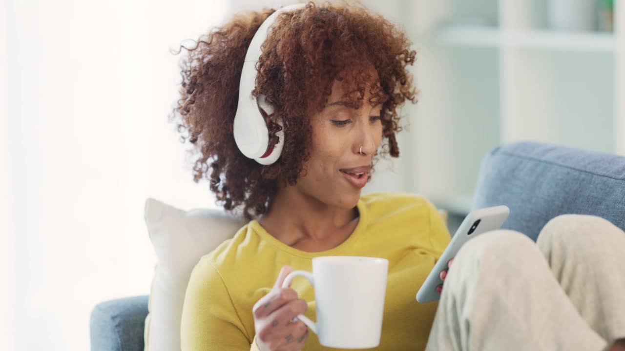 快乐的女人在家边喝咖啡边听音乐和唱歌。欢快的年轻女性干扰她最喜欢的歌曲，享受一个无忧无虑的周末，放松在沙发上，心情很好视频素材